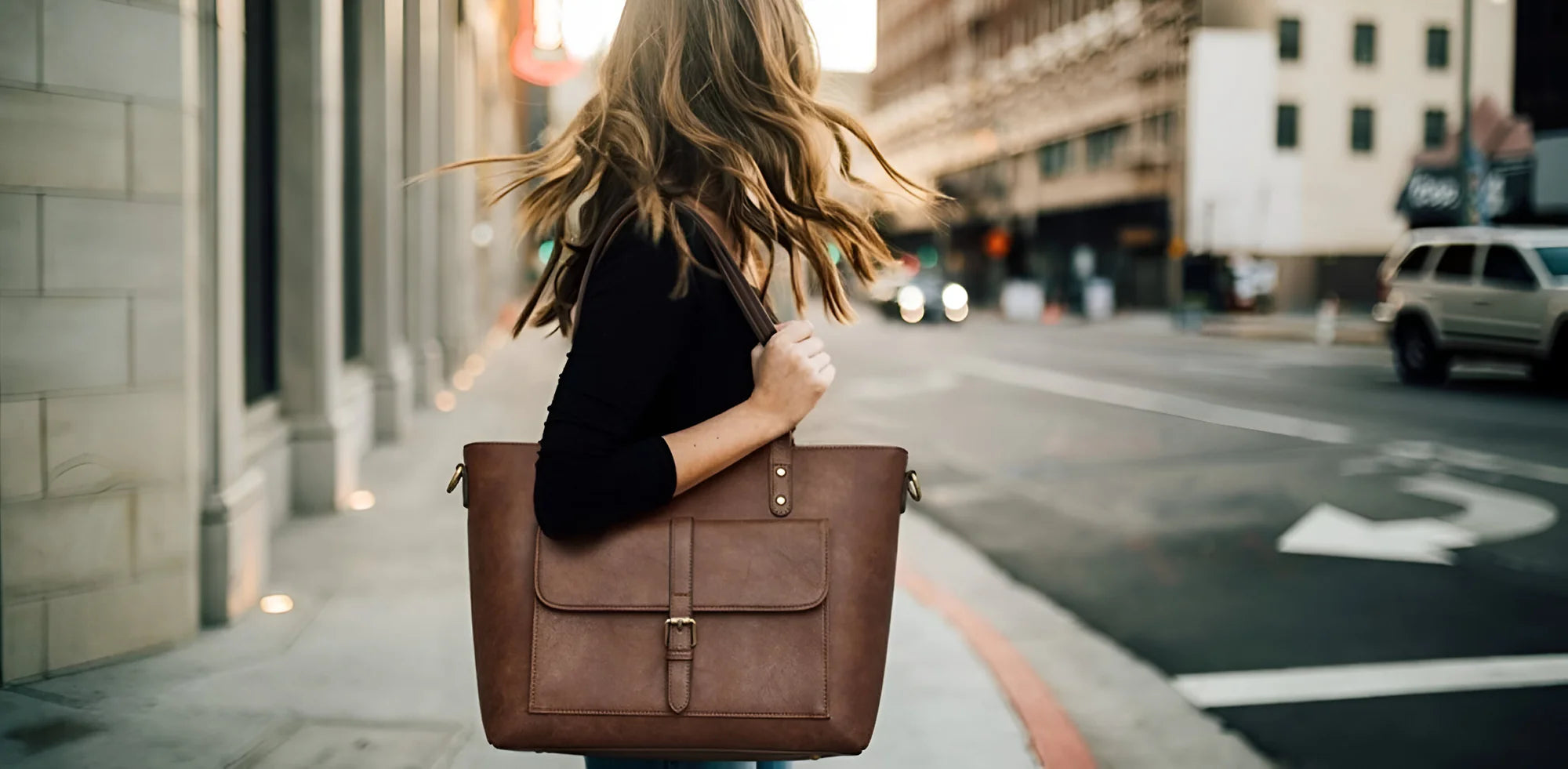 A woman with wavy hair walks on a city street, carrying a large brown tote bag. The background shows buildings and a blurred car, conveying a lively urban scene.