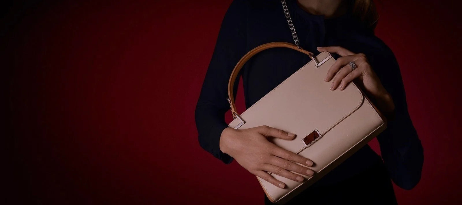 A person holds an elegant beige handbag with a brown handle in front of a deep red background.